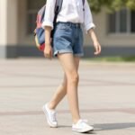 Girl wearing high-waisted denim shorts with a lightweight white button-down shirt and canvas sneakers for summer school outfit ideas