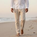 Man wearing beige linen pants with a white linen shirt walking on a beach at sunset, showcasing the perfect casual linen pants outfit for men