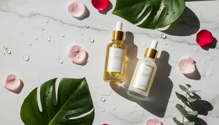 Luxurious serum bottles with droppers on marble surface surrounded by fresh botanicals and water droplets